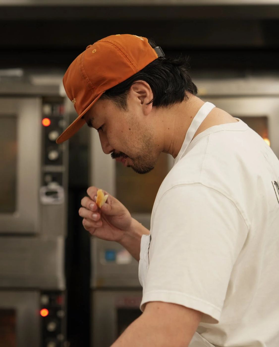 Hisashi Murakami in an orange cap, tasting a piece of bread by the ovens